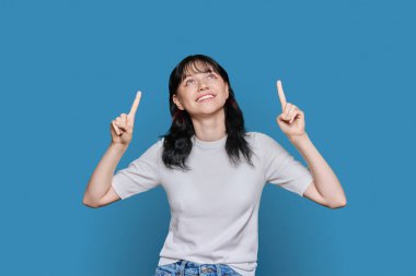 Parmakla işaret eden genç bir kadın, metin için yer, mavi arkaplan, yukarı bakan çekici genç kız. Reklamcılık, pazarlama konsepti