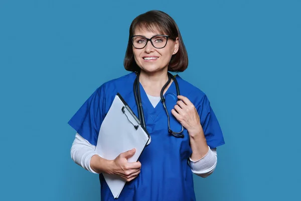 Portrait of female doctor with clipboard looking at camera on blue color background. Middle aged medic nurse with stethoscope, wearing blue uniform. Medicine, health care, staff concept