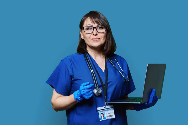 Portrait of serious female doctor with laptop, confident female looking at camera on blue studio background. Medic nurse with stethoscope in blue uniform. Health care medicine internet service staff