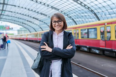 Tren istasyonunda sırt çantalı bir kadın bekliyor. Kollarını kavuşturup kameraya bakan bir kadın. Demiryolu taşımacılığı, yolcu taşımacılığı, yolculuk, seyahat, seyahat, insanlar