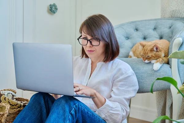 Middle aged woman using laptop along with sleeping cat lying on armchair at home, female using laptop for leisure and work. Home, job, freelance, lifestyle, pet, 40s people concept