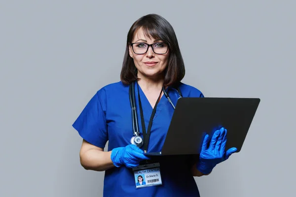 Friendly female doctor with laptop looking at camera on grey studio background. Smiling female in blue uniform with stethoscope, ID card posing in studio. Health care medicine internet service staff
