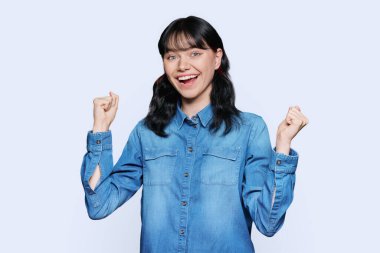 Mutluluk hissi, iyi şans, zafer, kutlama, beyaz stüdyo arka planında ağzı açık, yumruklarını kaldıran genç mutlu kadın. Çekici genç kız öğrenci kameraya bakarak eğleniyor.