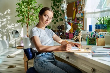Masada oturmuş kameraya bakan genç bir öğrencinin portresi. Gülümseyen yaratıcı kız 15-16 yaşlarında, kalem çizimleriyle. Eğitim, yaratıcılık, eğlence, hobi, gençlik kavramı.
