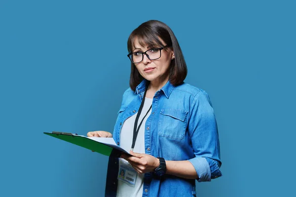 Portrait of woman worker with name identity card of industrial center clipboard, on blue color background. Confident female engineer manager supervisor auditor inspector expert looking at camera