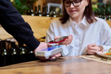 Woman in restaurant sitting at table with food paying for order using smartphone and wireless bank payment terminal in hands of waiter. Cashless technology, money service, leisure, lifestyle, people