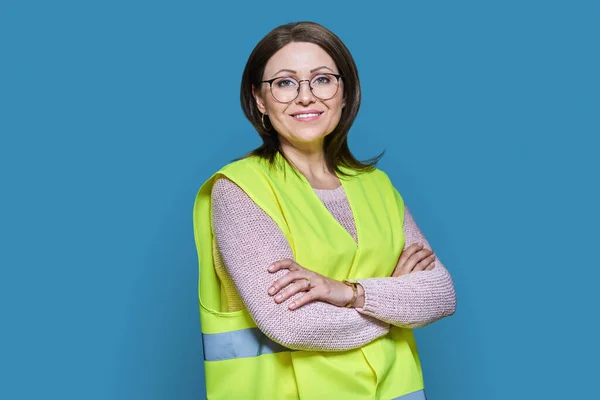Portrait of female industrial construction worker in vest looking at camera with crossed arms on blue background. Logistics construction industry management architecture engineering staff concept