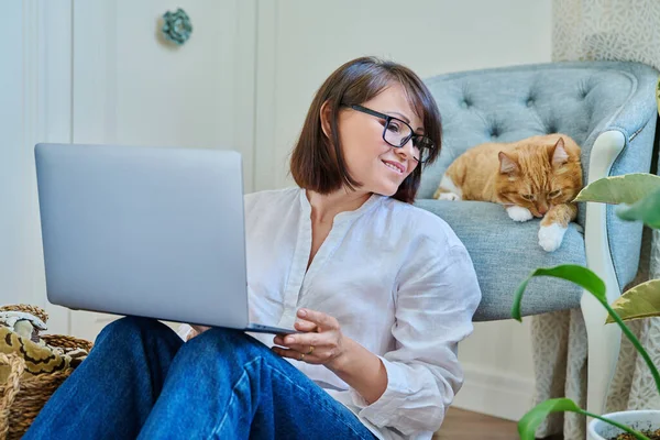 Middle aged woman using laptop along with sleeping cat lying on armchair at home, female using laptop for leisure and work. Home, job, freelance, lifestyle, pet, 40s people concept