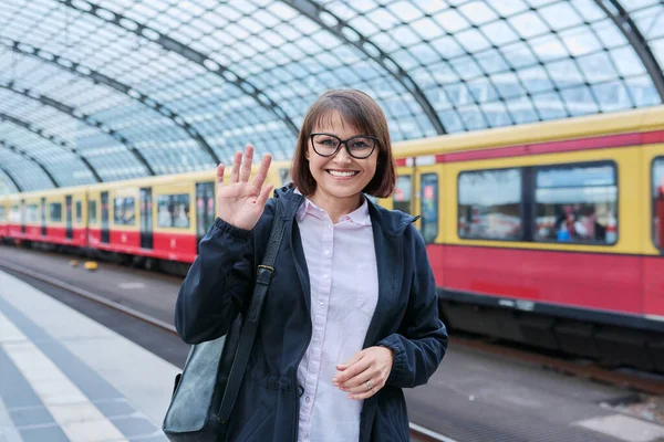 Woman passenger with backpack waiting on railway platform inside station, smiling female with crossed arms looking at camera. Rail transport, passenger transportation, journey, travel, trip, people