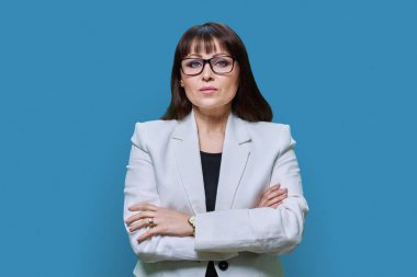 Confident serious middle aged business woman in white jacket on blue studio background. Businesswoman with crossed arms looking at camera. Leadership management teaching finance banker businessperson