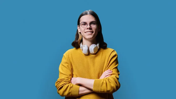 Confident young male posing with crossed arms against blue color studio background. Smiling cheerful student, guy 18, 19 years old looking at camera. Youth, age, fashion, style, lifestyle concept