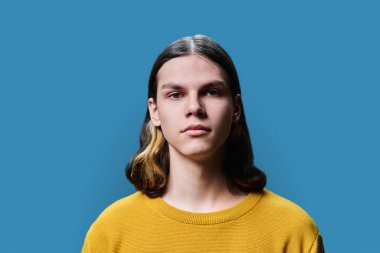 Headshot portrait of a serious teenage guy 18, 19 years old, on blue color studio background. Confident handsome young male looking at camera