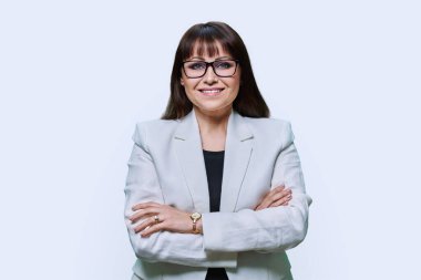 Hafif stüdyo arka planında beyaz takım elbiseli, orta yaşlı, kendine güvenen bir iş kadını. Kollarını kavuşturup kameraya bakan bir iş kadını. Liderlik yönetimi finans bankacısı iş adamı