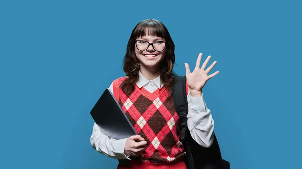 Portrait of smiling joyful female student with laptop backpack waving hand, on blue studio background. Cheerful delightful girl in glasses looking at camera. College university courses, education