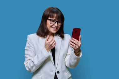 Middle aged beautiful businesswoman with phone in hands on blue studio background. Smiling mature female in jacket, glasses, looking at smartphone. Technology business lifestyle businessperson people