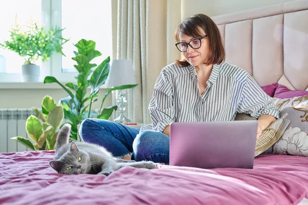 Middle aged woman at home on bed with laptop and cat. Mature 40s female looking at screen, on sofa with pet. Work at home, technology, leisure, freelancing, lifestyle, people and animals concept