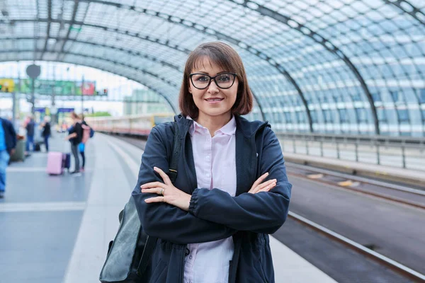 Woman passenger with backpack waiting on railway platform inside station, smiling female with crossed arms looking at camera. Rail transport, passenger transportation, journey, travel, trip, people