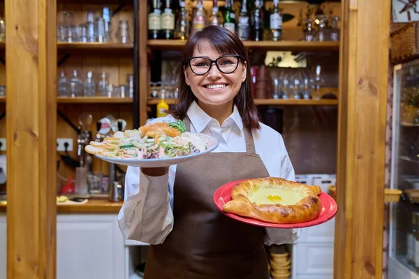 Middle aged female restaurant owner manager administrator posing in an apron with plates of cooked food. Small business, entrepreneurship, cafe cafeteria restaurant, service, job success concept