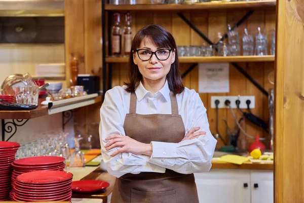 Portrait of middle aged woman bistro restaurant manager owner in an apron, confident smiling looking at camera with crossed arms. Small business, service, work, staff concept
