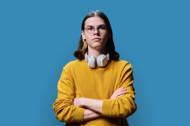 Confident young male posing with crossed arms against blue color studio background. Serious handsome student, guy 18, 19 years old looking at camera. Youth, age, fashion, style, lifestyle concept