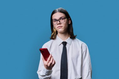 Young business male using smartphone on blue studio background. Serious guy in shirt with tie looking at camera. Mobile applications on smartphone for education, business, work, leisure, communication