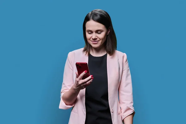 Young business woman using smartphone on blue color background. Positive smiling female in glasses jacket looking texting on phone. Mobile applications, digital technologies in business work leisure