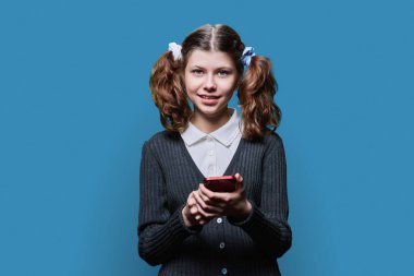 Preteen smiling school girl with smartphone in hands looking at camera on blue studio background. Texting messaging, use of mobile phone for study and leisure, lifestyle technology childhood concept