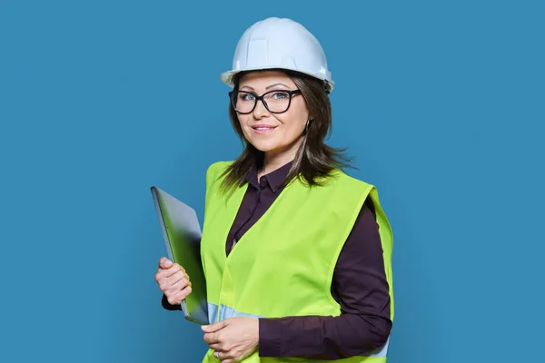 Portrait of engineer manager woman in helmet vest holding laptop, over blue studio background. Logistics, construction, industry, management, architecture, engineering, staff workers concept