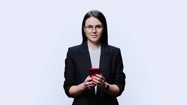Young business woman using smartphone on white studio background. Positive smiling female in glasses black suit looking at camera. Mobile applications digital technologies in business work leisure