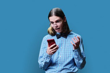 Mavi stüdyo arka planında akıllı telefondan bakan neşeli, yakışıklı genç bir erkek. Şok olmuş gözlüklü, papyonlu zeki çocuk, iyi haber, sürpriz mutluluk konsepti.