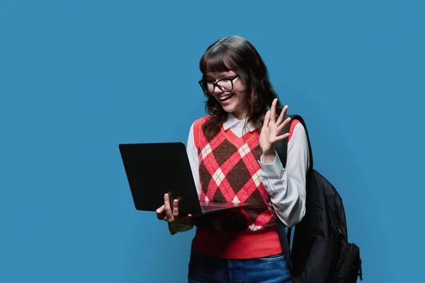 Smiling female student 18, 19 years old looking into laptop on blue studio background. Cheerful girl in glasses using internet for study, e-learning, online lessons, college university courses