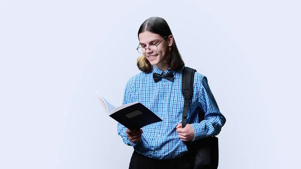 Portrait of smiling young male college student, reading notebook on white studio background. Teenage guy in glasses with backpack looking at book, smart handsome dressed in shirt with bow tie