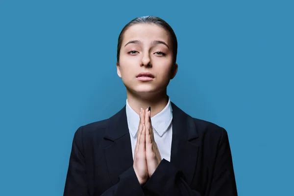 Young female in glasses pleading holding her palms together, on blue studio background. Girl student looking at camera, in request, in plea, asking for forgiveness for help sorry pray please