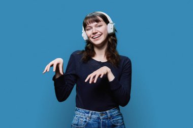 Cheerful attractive young female in headphones dancing trendy dance, on blue color studio background. Modern youth, fashion, music, dancing, fun, lifestyle, people concept