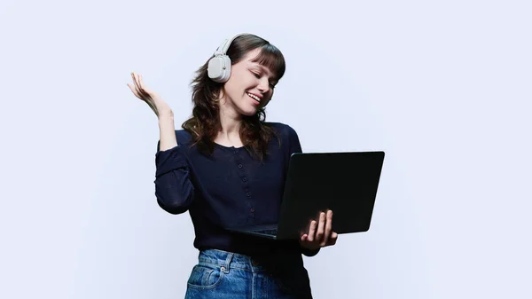 快乐的年轻女性在耳机里看着笔记本电脑 白色的工作室背景 情感快乐迷人的女孩上网聊天 听唱歌 休息和休闲 科技生活方式的概念 — 图库照片