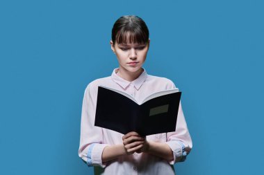 College university student reading exercise book, on blue color studio background. Serious focused girl looking at notebook, education knowledge learning concept