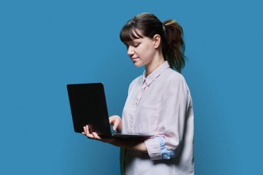 Serious concentrated female student looking at laptop screen, on blue studio background. Smart intelligent girl using internet for study e-learning online lessons college university courses