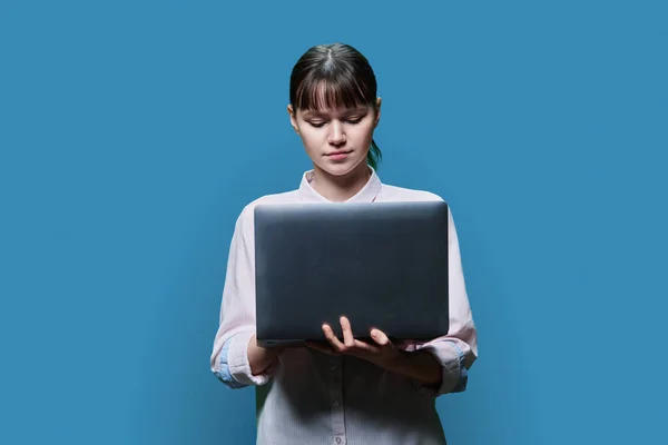 Serious concentrated female student looking at laptop screen, on blue studio background. Smart intelligent girl using internet for study e-learning online lessons college university courses