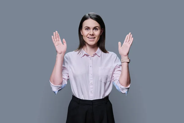 Happy surprised young woman looking at camera on gray studio background. Joyful emotional female 30 years old in shirt with raised hands. Expression, shock, surprise, joy, youth people concept