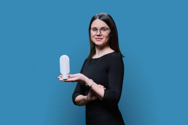 Young woman holding white clean tube of body face cream, on blue studio background. Advertising sale of cosmetics for care, treatment, dermatology, cosmetology, beauty industry concept