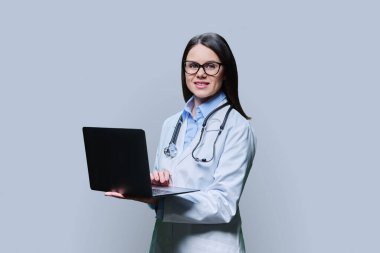 Young friendly female doctor using laptop computer on light gray studio background. Confident female in white coat with glasses looking at camera. Medical staff occupation health care science medicine