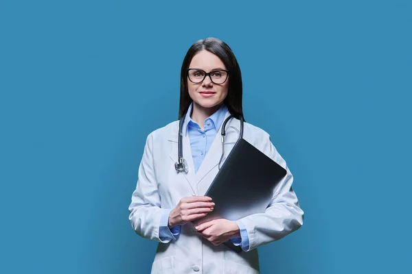 Young friendly female doctor with laptop computer on blue color studio background. Confident female in white coat with glasses looking at camera. Medical staff occupation health care science medicine