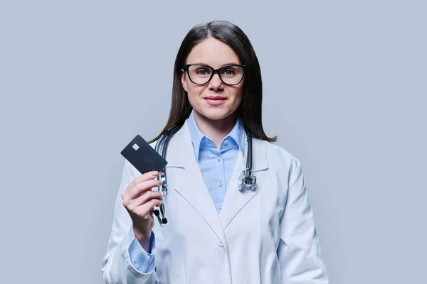 Young woman doctor holding credit card on light gray studio background. Friendly female looking in camera. Insurance, healthcare, credit card service concept