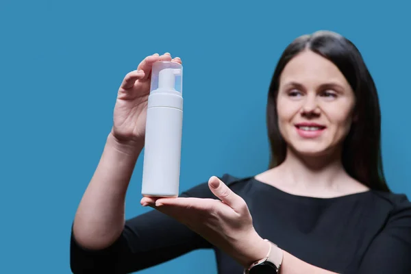 Close-up of white clean tube with cosmetic beauty product in hands of woman on blue background. Professional medical care cosmetics for face body hair, gel foam for washing, cosmetic milk lotion cream