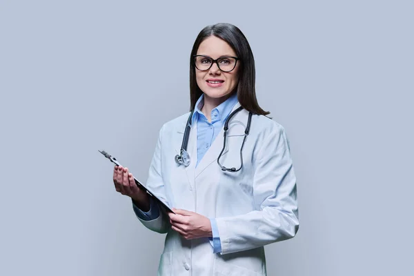 Young friendly woman doctor with clipboard on light gray studio background. Confident female in white coat with glasses looking at camera. Medical staff occupation health care science medicine concept