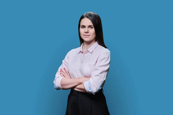 Young smiling woman with crossed arms looking at camera on blue color studio background. Beautiful confident successful. Young people 30 years old, business work, lifestyle concept