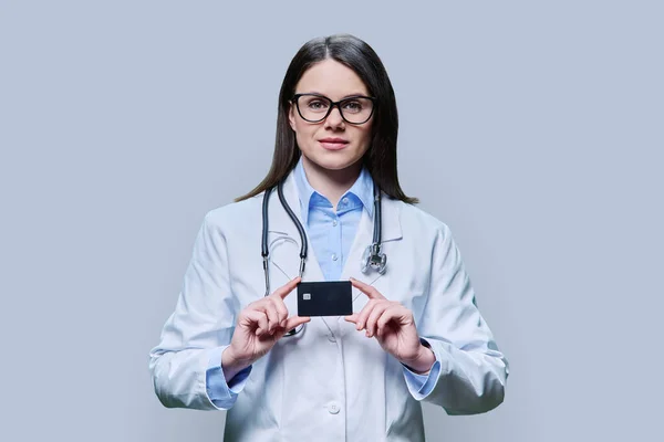 Young woman doctor holding credit card on light gray studio background. Friendly female looking in camera. Insurance, healthcare, medicine, credit card service concept