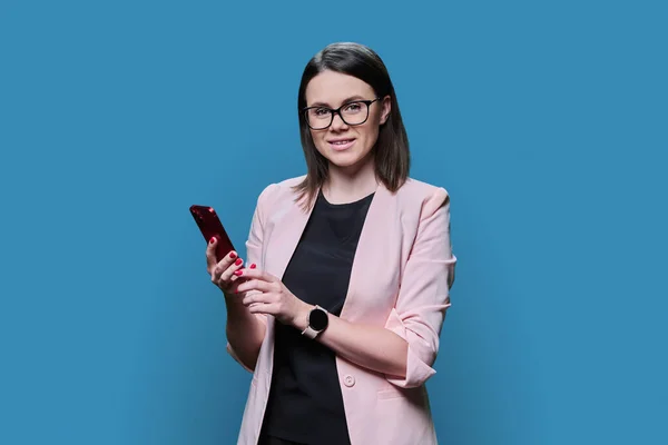 Young business woman using smartphone on blue color background. Positive smiling female in glasses jacket looking at camera, texting. Mobile applications, digital technologies in business work leisure