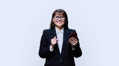 Gülümseyen orta yaşlı iş kadını akıllı telefon kullanıyor, beyaz stüdyo geçmişi var. Siyah takım elbiseli başarılı bir kadın kameraya bakıyor. İş, yönetim, iş, mobil uygulamalar dijital teknolojiler