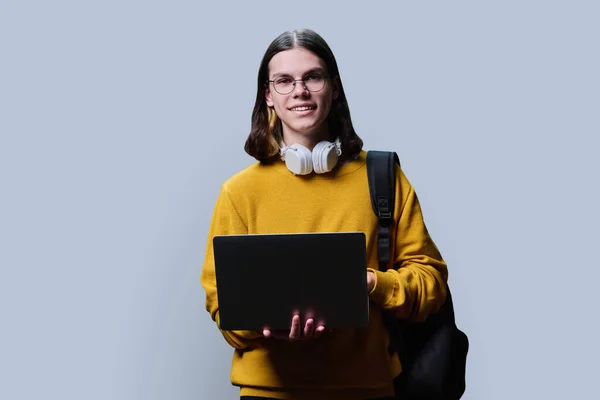 Portrait of young male college student using laptop backpack on grey studio background. Handsome trendy guy in glasses headphones looking at camera. Youth, education, high school college university